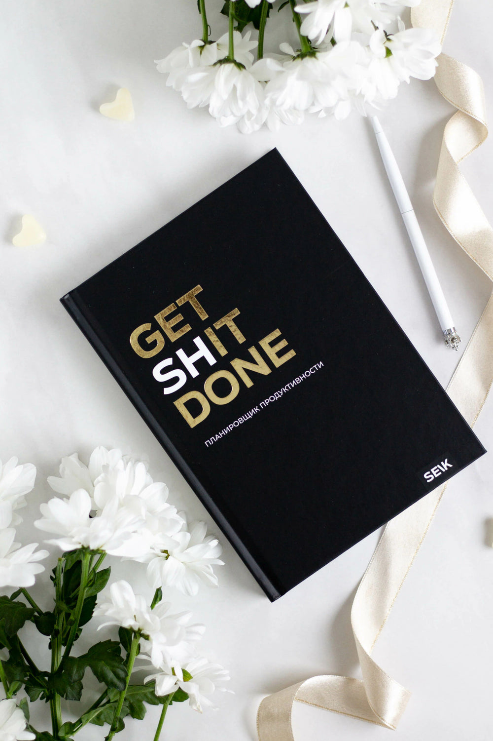 Чёрный планировщик Get Shit Done среди белых цветов, ленты и ручки.