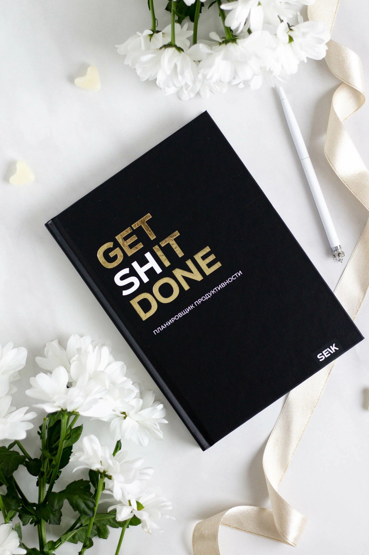 Чёрный планировщик Get Shit Done среди белых цветов, ленты и ручки.