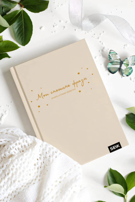 Книга «Мои смешные фразы» на белом фоне с листьями, бабочкой и вязаной тканью